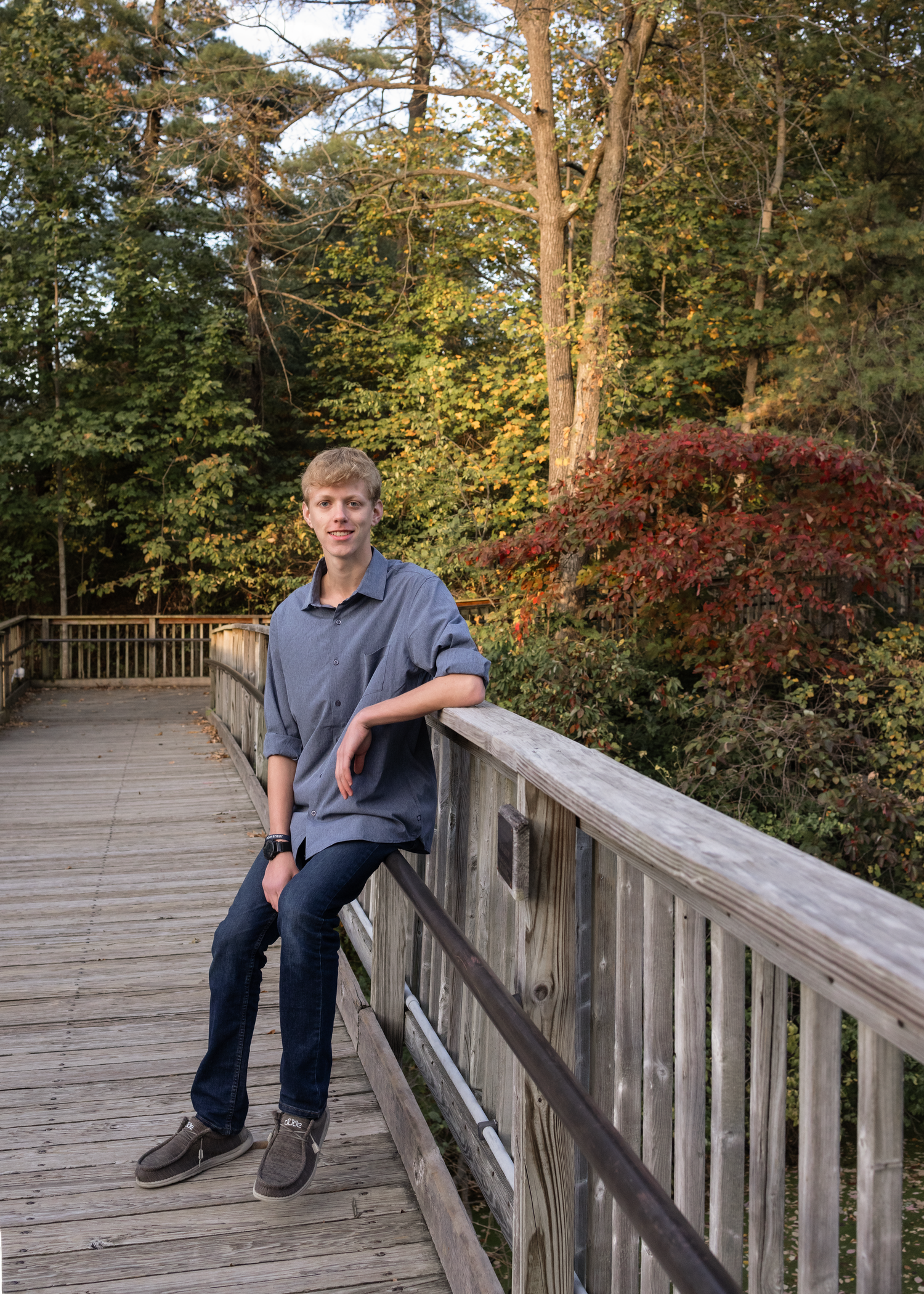 A person sits casually on a wooden boardwalk railing surrounded by autumn foliage in a peaceful forest setting.