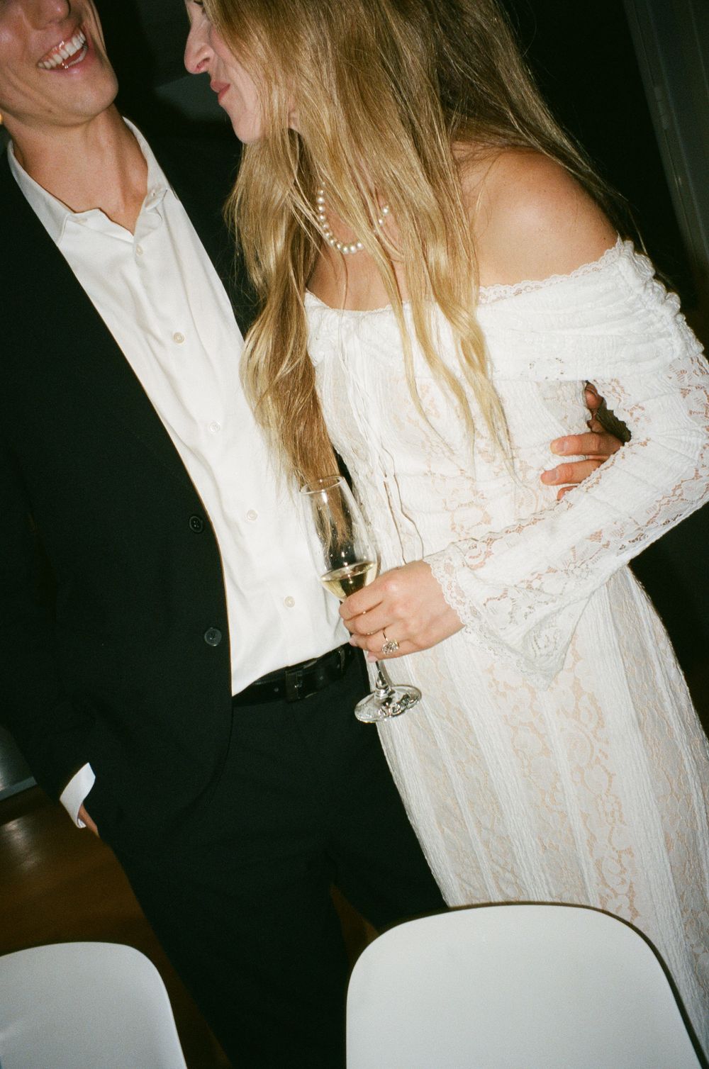 A couple in formal attire embraces at a celebration, with the bride wearing a white lace gown and holding a champagne glass.