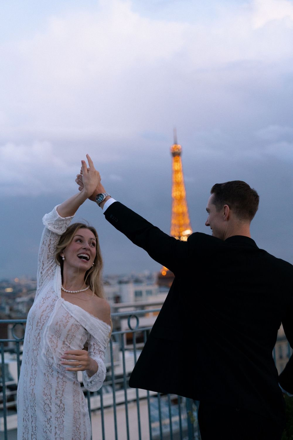 A couple dances together on a rooftop terrace with the illuminated Eiffel Tower glowing in the twilight background.