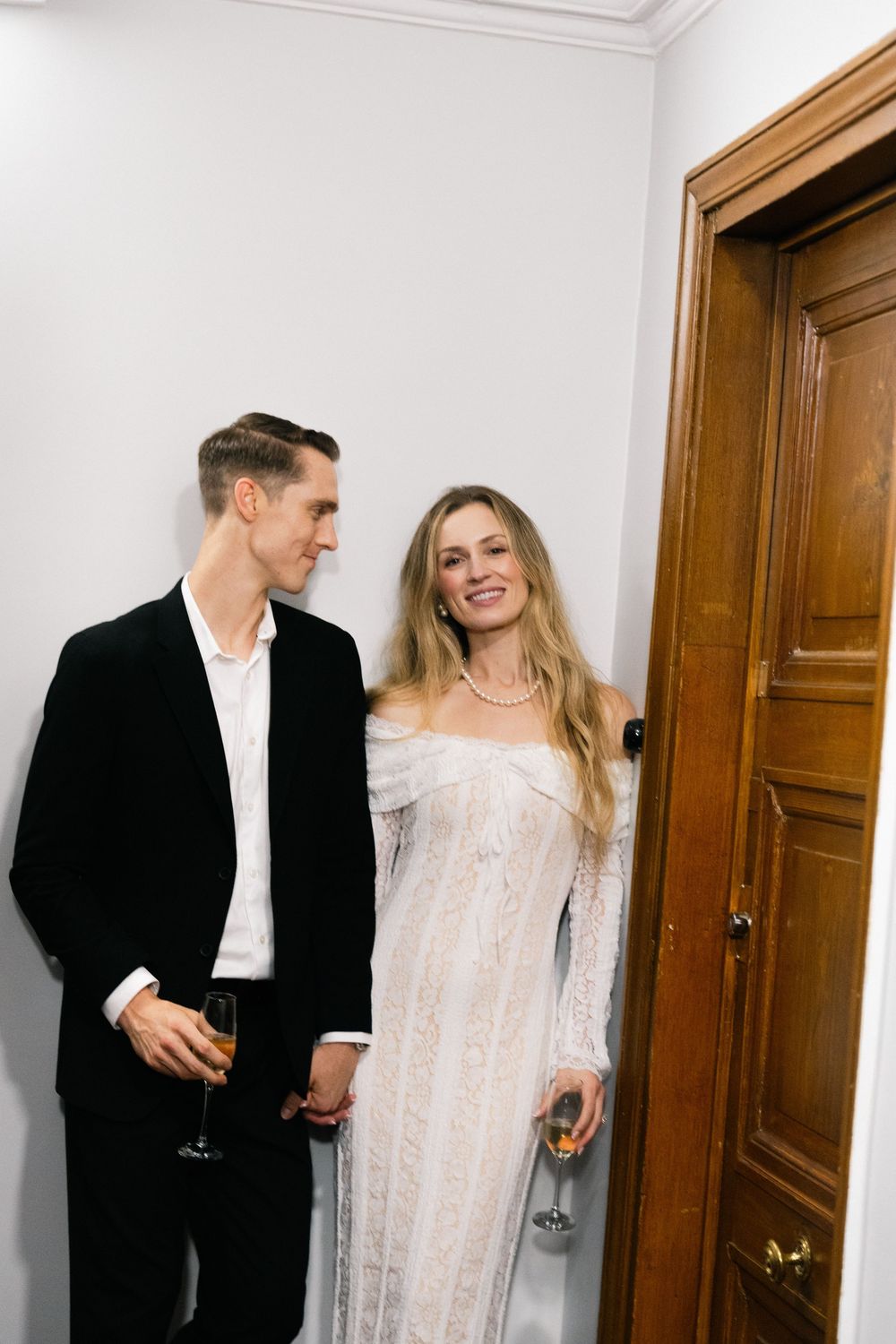 A couple in formal attire shares an intimate moment in a hallway with white walls and wooden doors.
