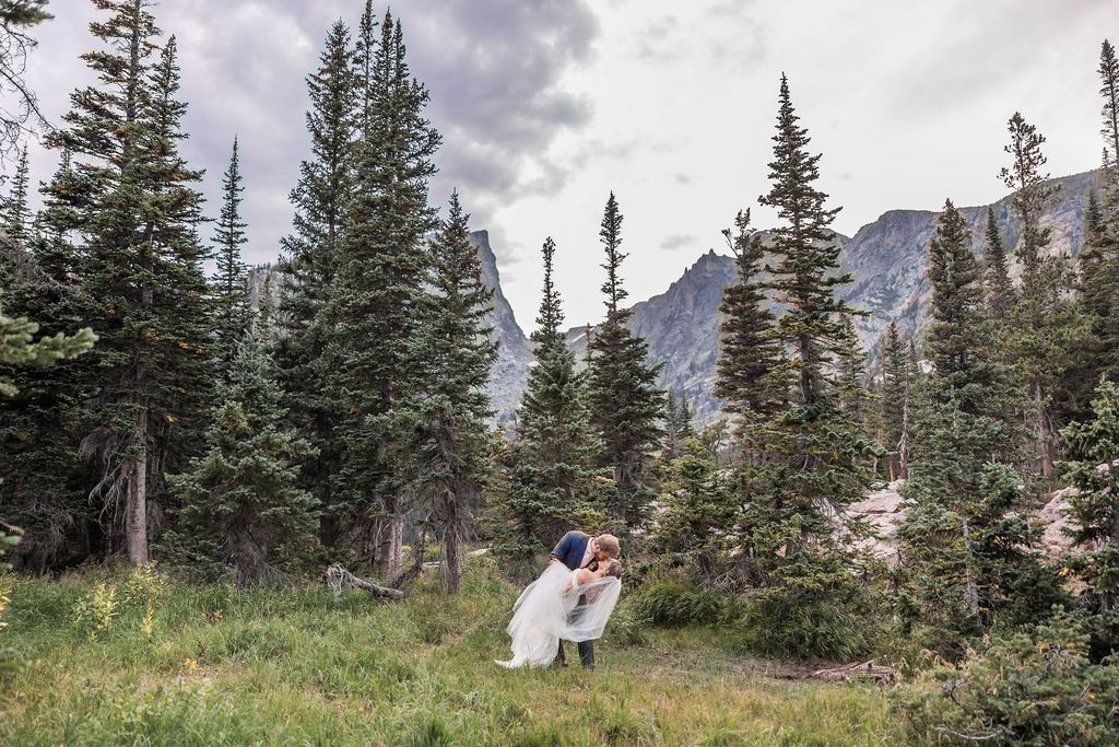 A couple embraces in a scenic mountain meadow surrounded by evergreen trees and dramatic cloudy skies.