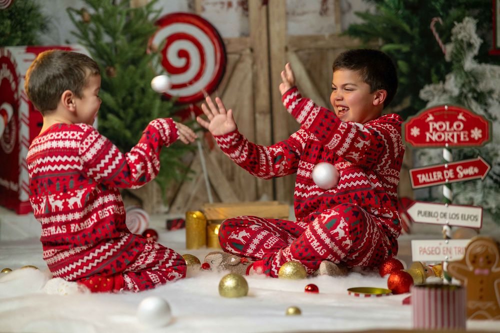 Children in matching red and white holiday pajamas play and laugh together on a wooden floor with Christmas decor.