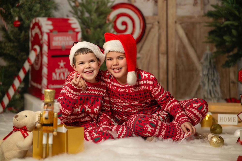 Children in festive red and white sleepwear sit and play near Christmas decorations and greenery.