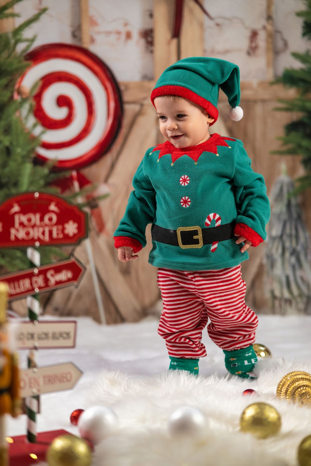 A baby wearing a festive green elf costume with red striped pants poses near Christmas decorations and North Pole signs.
