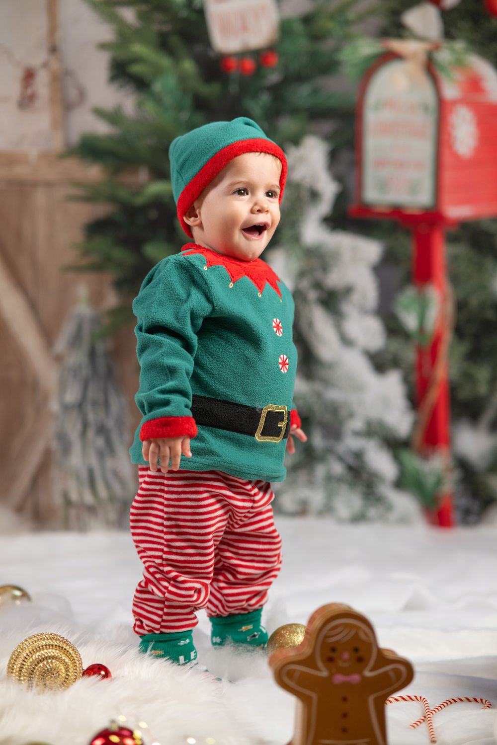 Baby in elf costume and striped pants plays in snowy Christmas scene with gingerbread decorations.