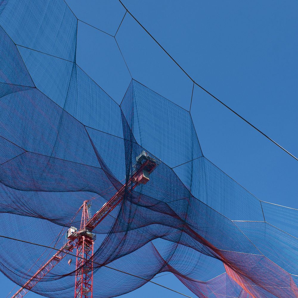 Blue construction netting creates abstract patterns against bright sky with red crane visible.