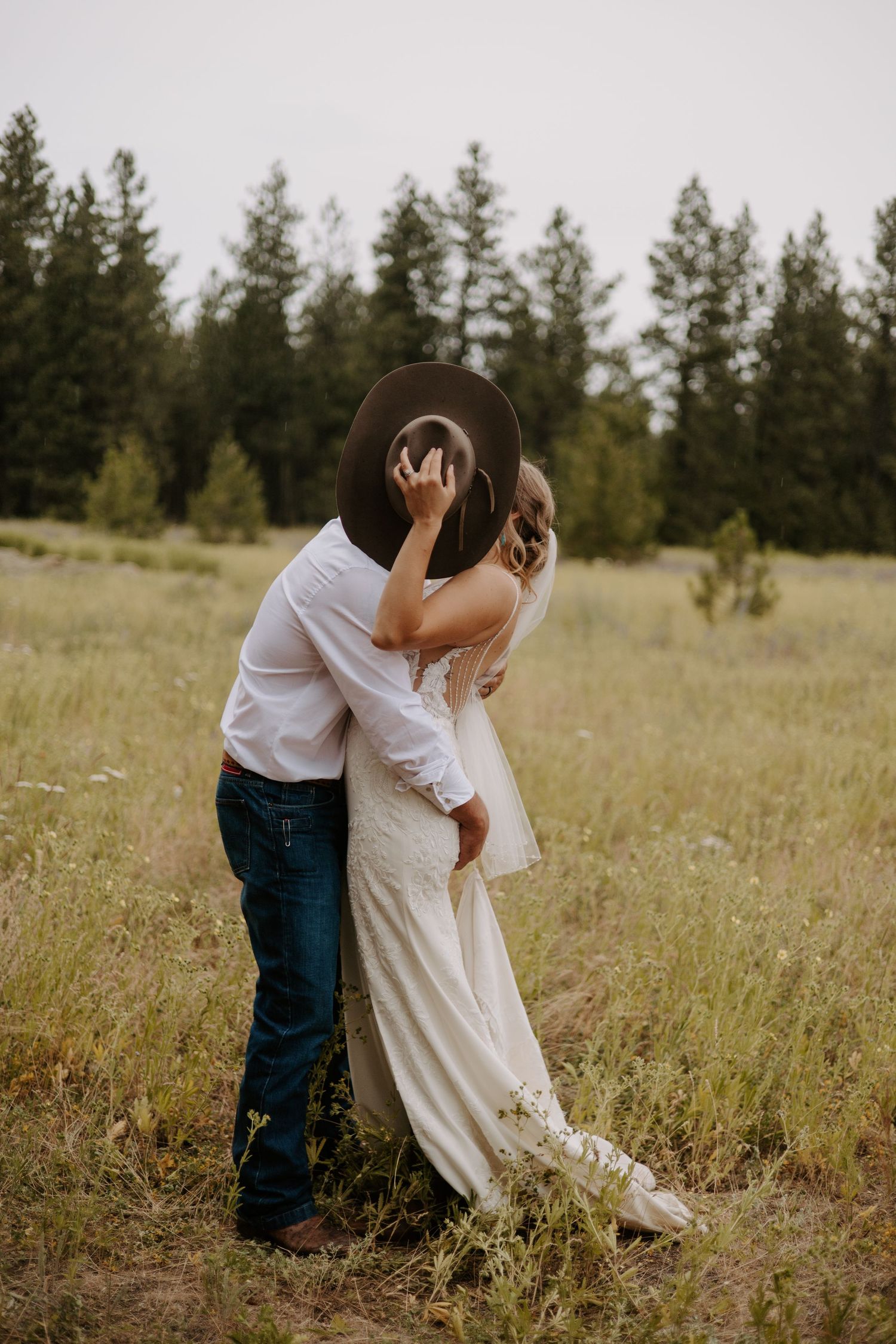 A couple embraces in a golden meadow with evergreen trees in the background during a romantic outdoor photoshoot.