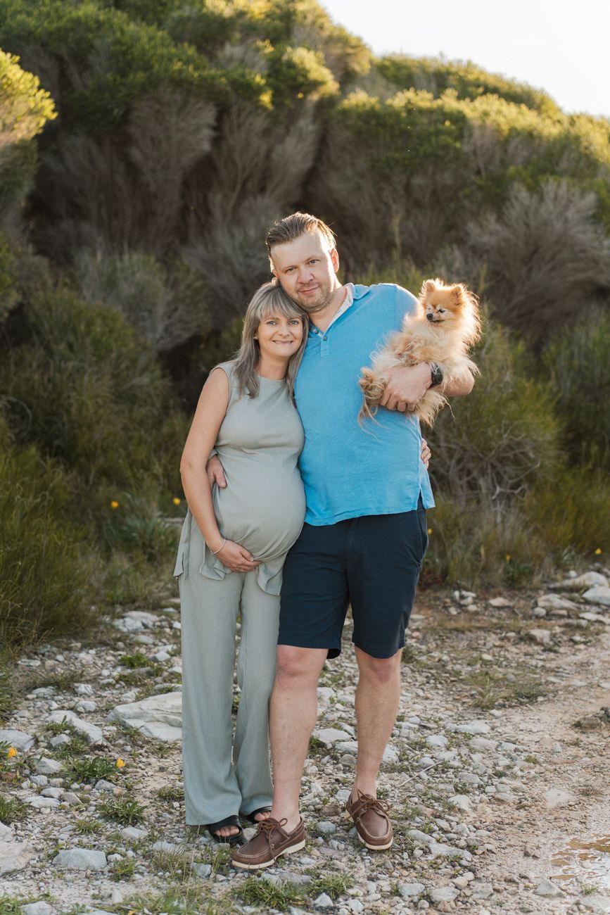 A couple and their small fluffy dog pose together on a rocky trail during golden hour maternity photoshoot in Maroubra Sydney