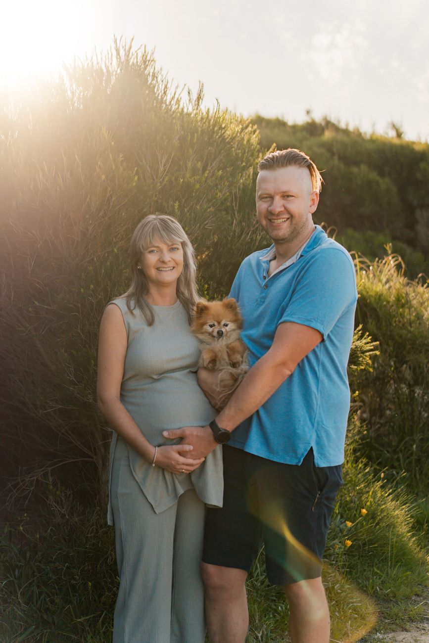 A couple poses with their dog pet in a sunlit outdoor setting during golden hour maternity photoshoot in Sydney Maroubra Beach