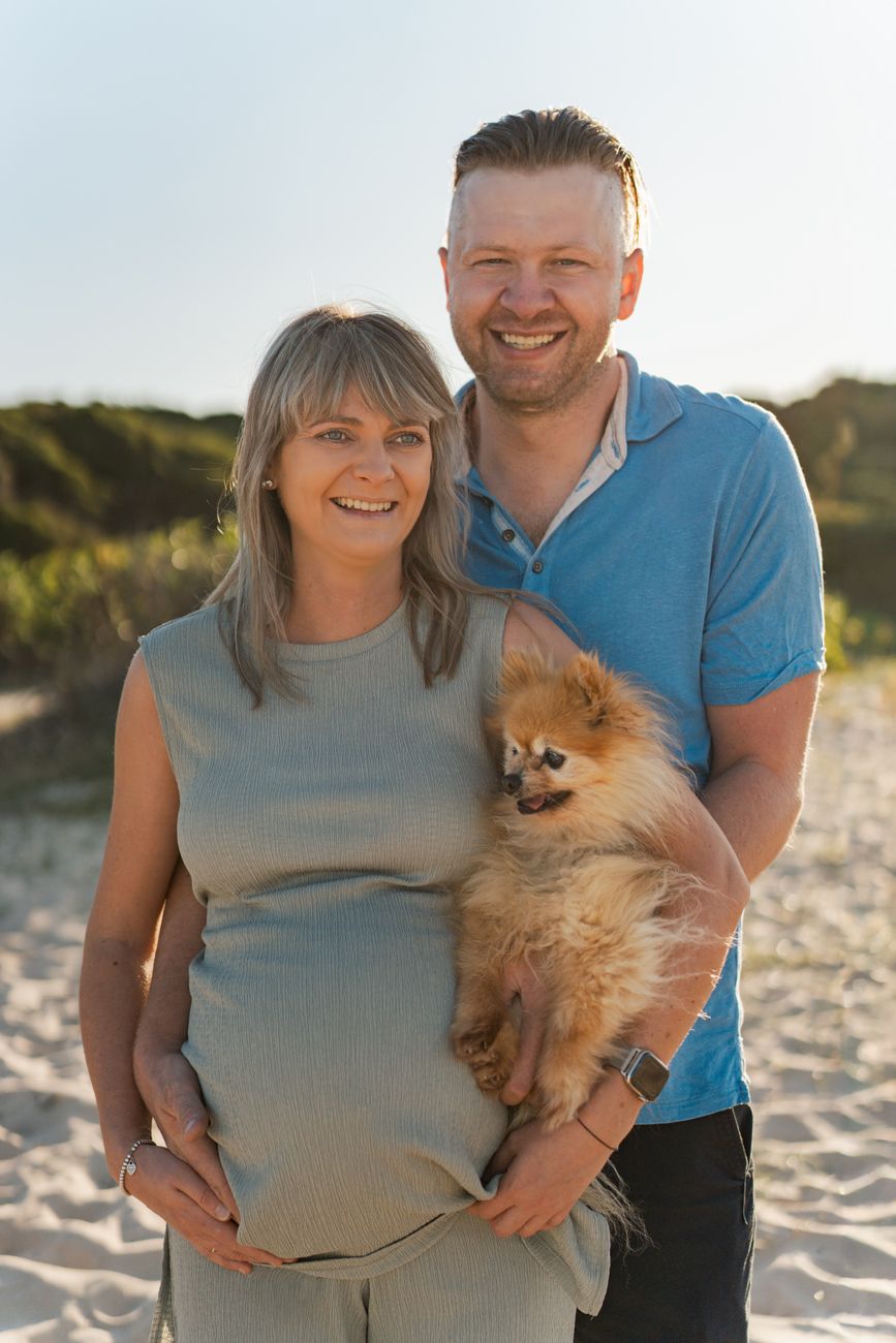 A couple poses on the beach with their fluffy Pomeranian dog during a sunny maternity photoshoot in Sydney Maroubra Beach