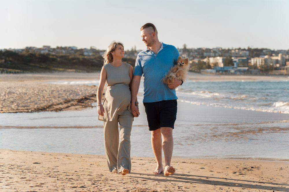 A couple walks along a sandy beach with their small dog while enjoying a sunny day by the ocean during their maternity photoshoot in Sydney