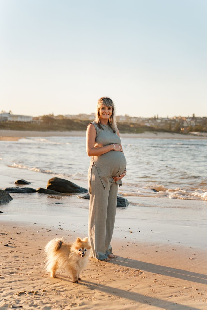 A pregnant woman stands along a sandy beach at sunset with a fluffy dog by her side during a maternity photoshoot in Sydney Maroubra Beach