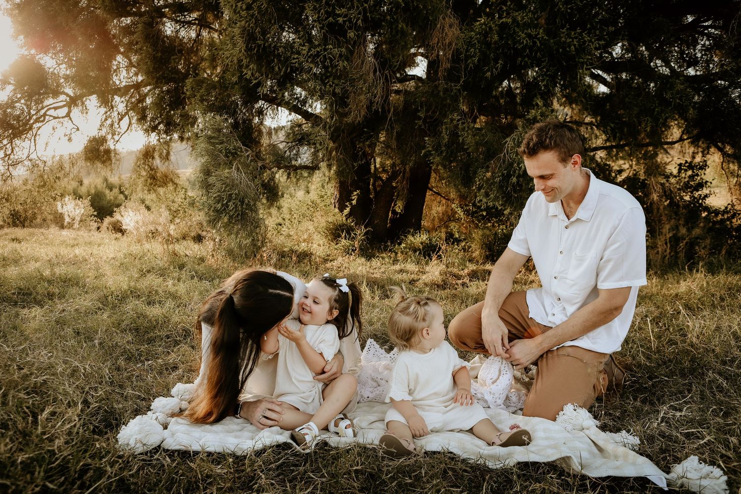 A romantic outdoor photo session on a white blanket during golden hour sunset in a rustic field setting.