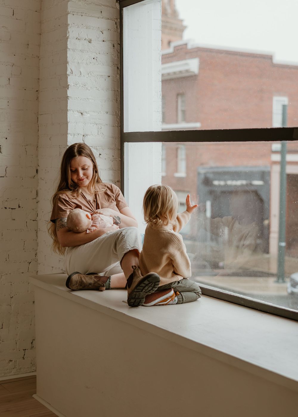 A parent and child sit together on a bright windowsill in a modern loft space with white brick walls.