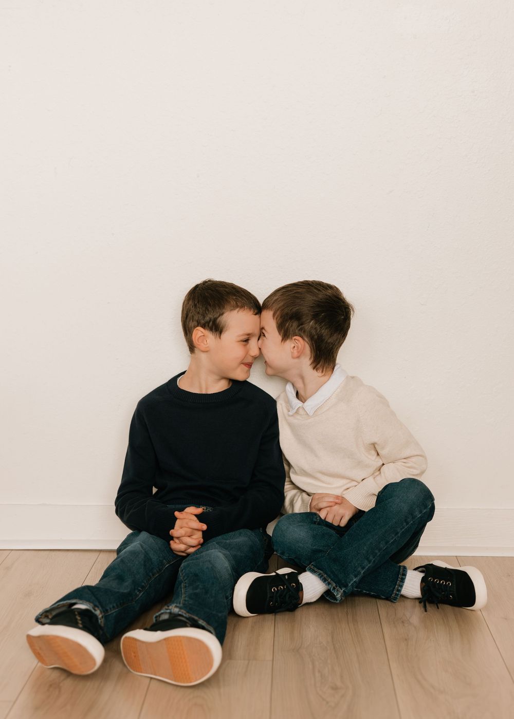 Two young siblings in casual wear sitting on wooden floor sharing playful moments in a minimalist setting.