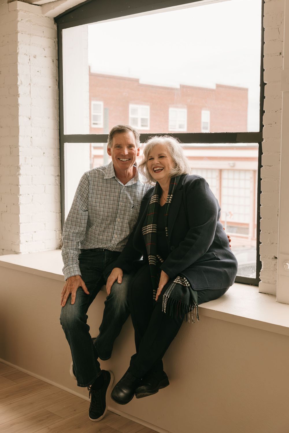 A couple sits together on a windowsill in a bright room with brick buildings visible through the window.