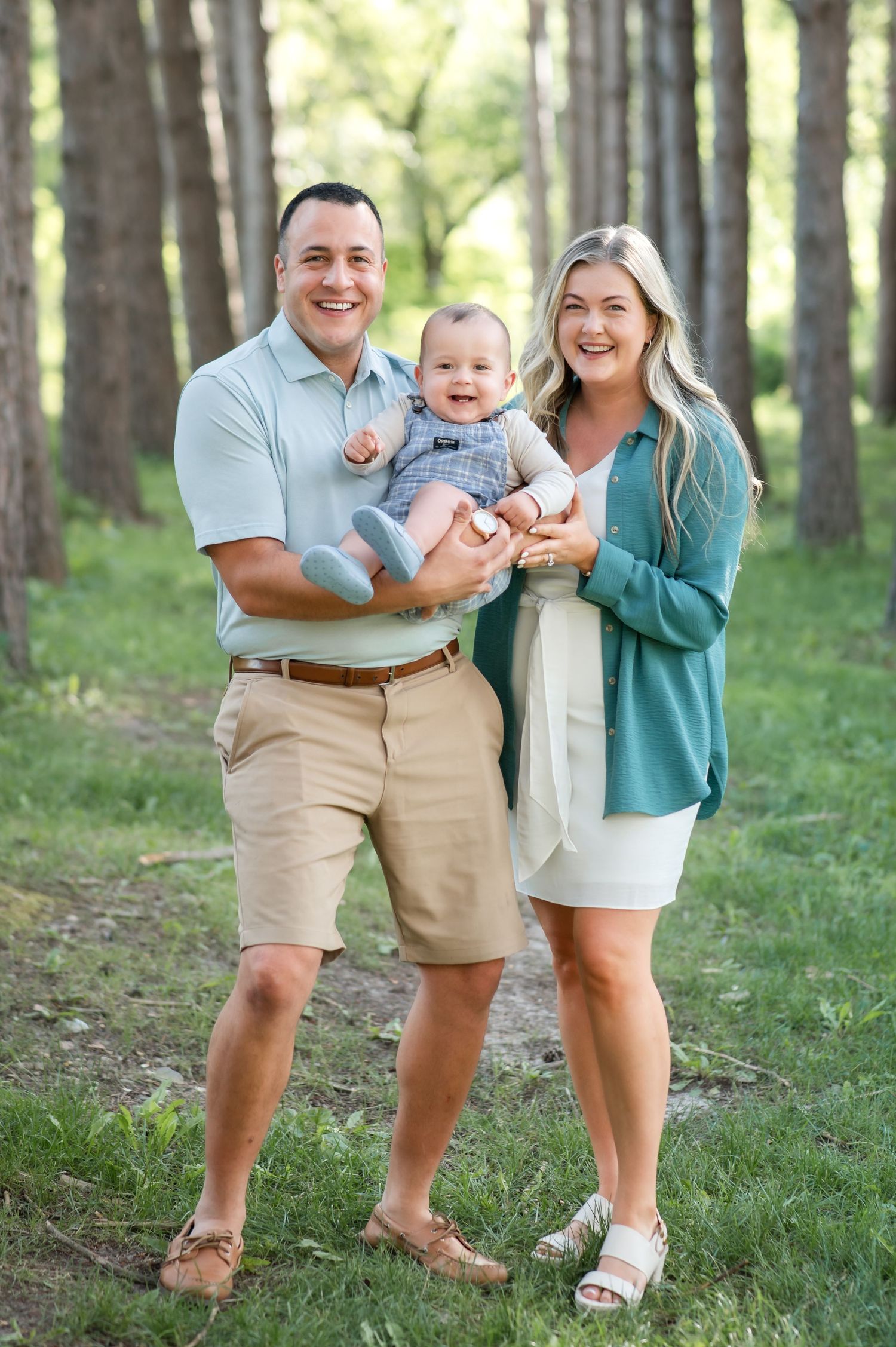 A family poses together in a sunlit forest setting wearing coordinated casual summer outfits.