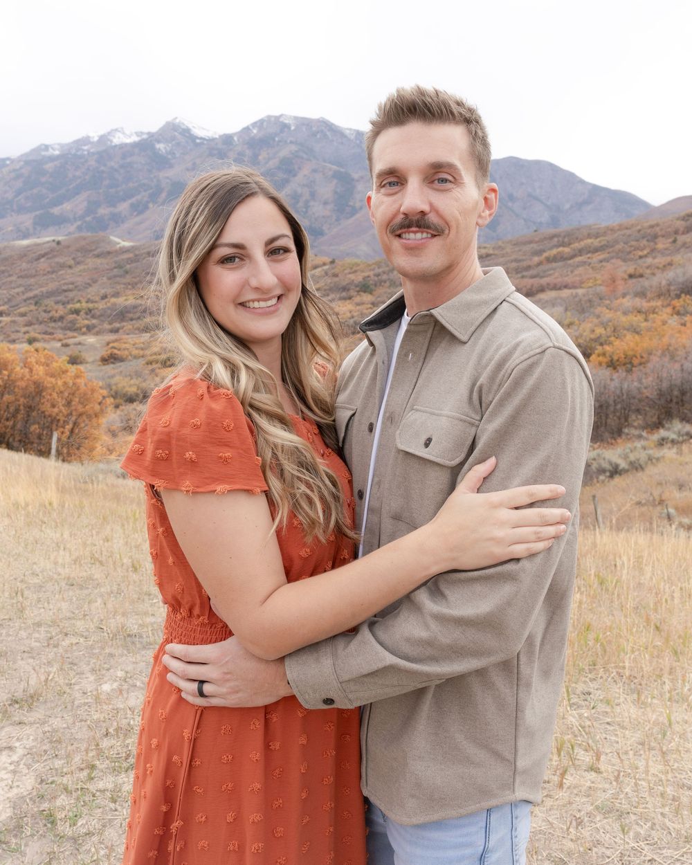 Couple embraces in a rustic autumn setting with mountains and golden grass in the background.