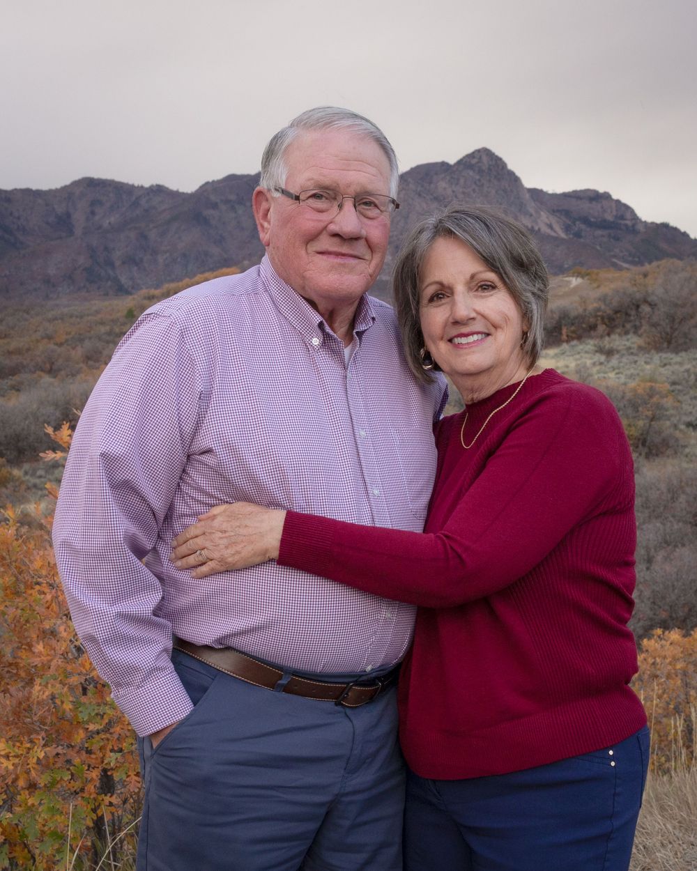 A couple stands together outdoors in front of rugged desert mountains on an overcast day.