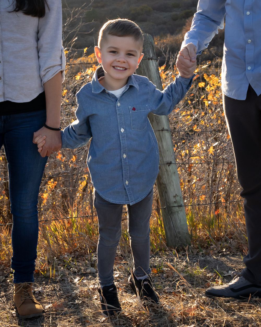 A family wearing denim and white shirts poses together outdoors during golden hour with sunlit foliage in the background.