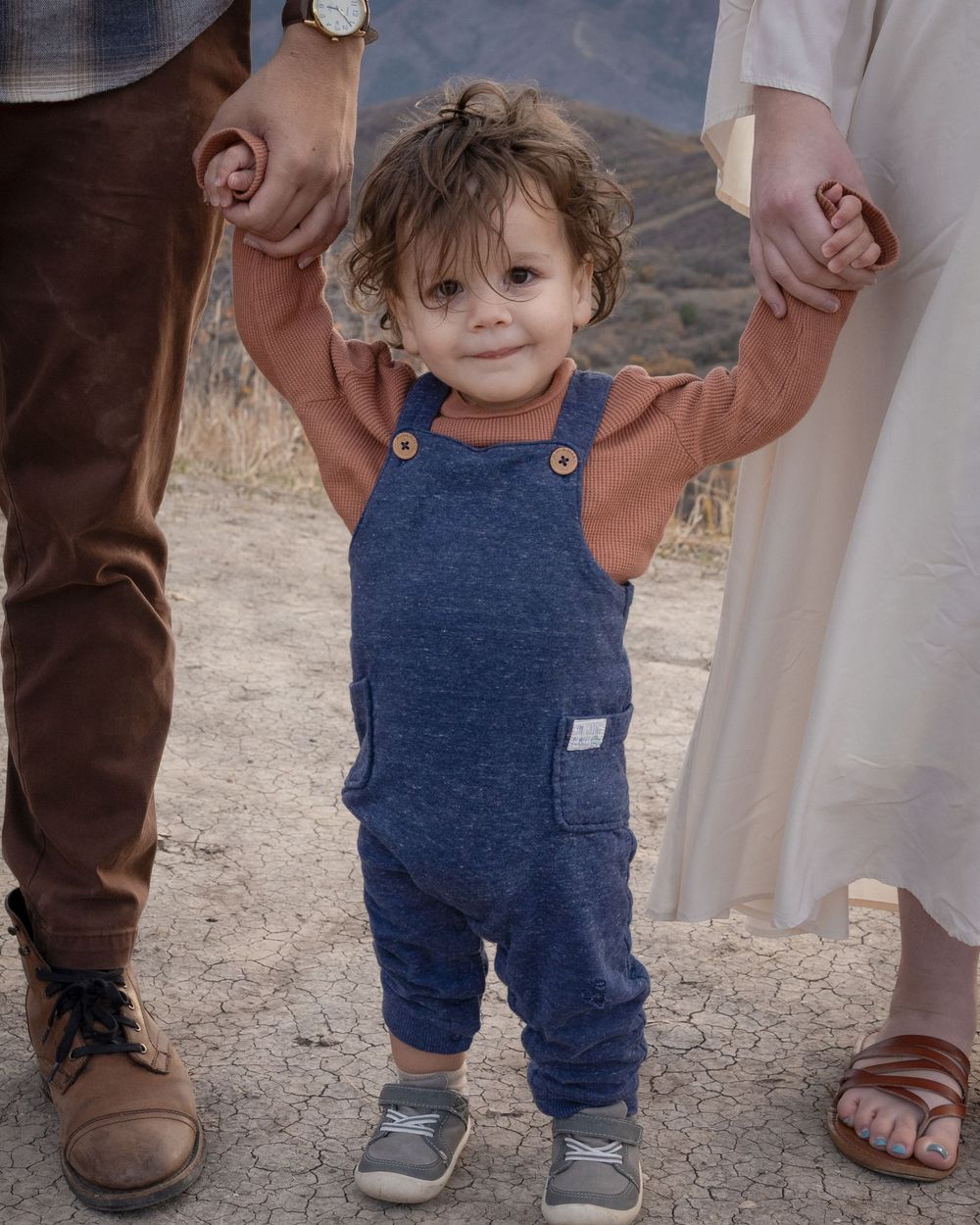 A small child in blue overalls holds hands with adults while taking steps on a dirt path.
