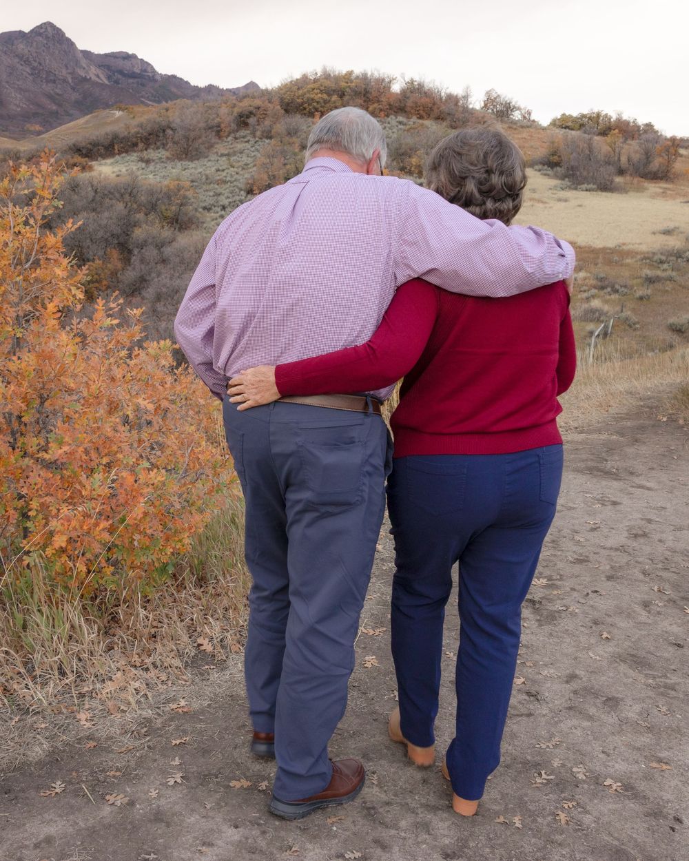 Two people walking together on a dirt trail surrounded by autumn foliage in a mountainous landscape.