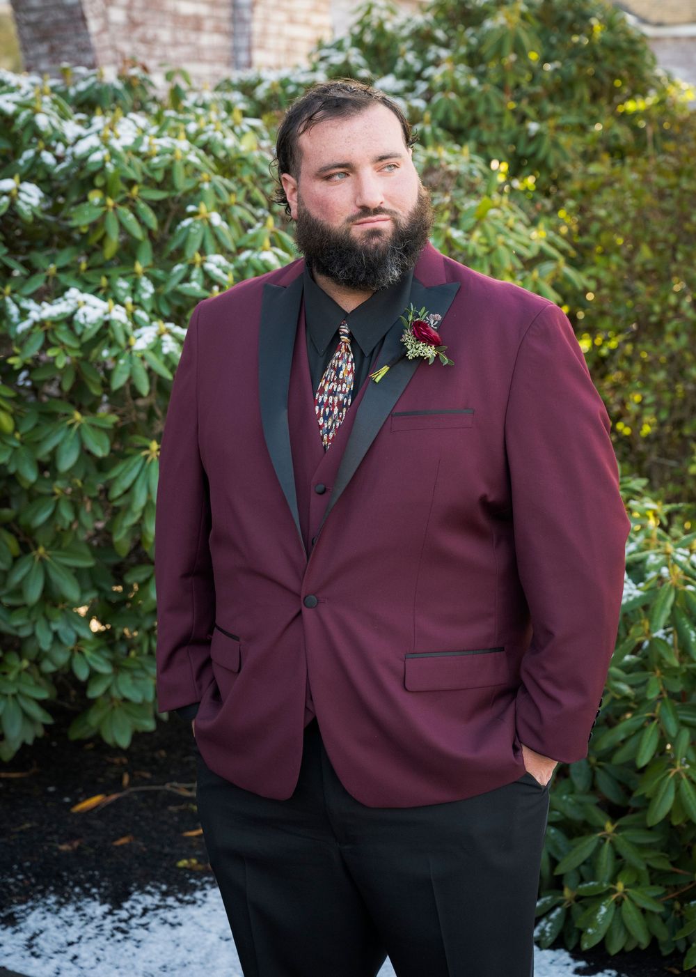 Groom in burgundy tuxedo jacket poses against garden backdrop with greenery.