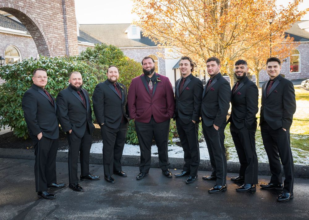 Groomsmen in formal black suits and burgundy jacket pose outdoors during autumn with fall foliage in background.