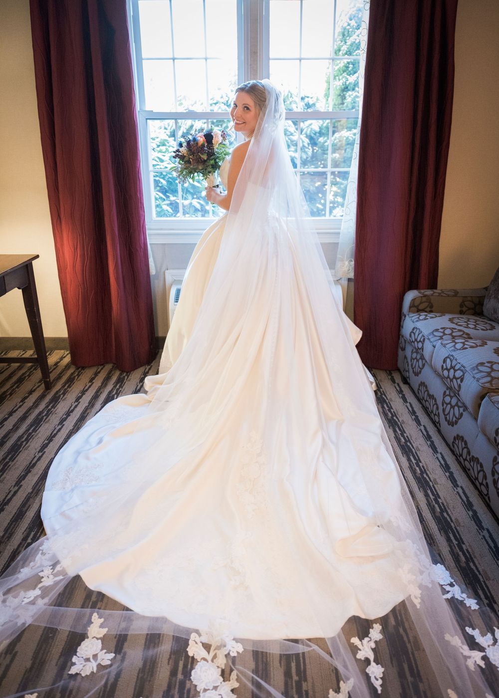 Elegant wedding dress with flowing train and veil displayed in a room with burgundy curtains and hardwood floors.