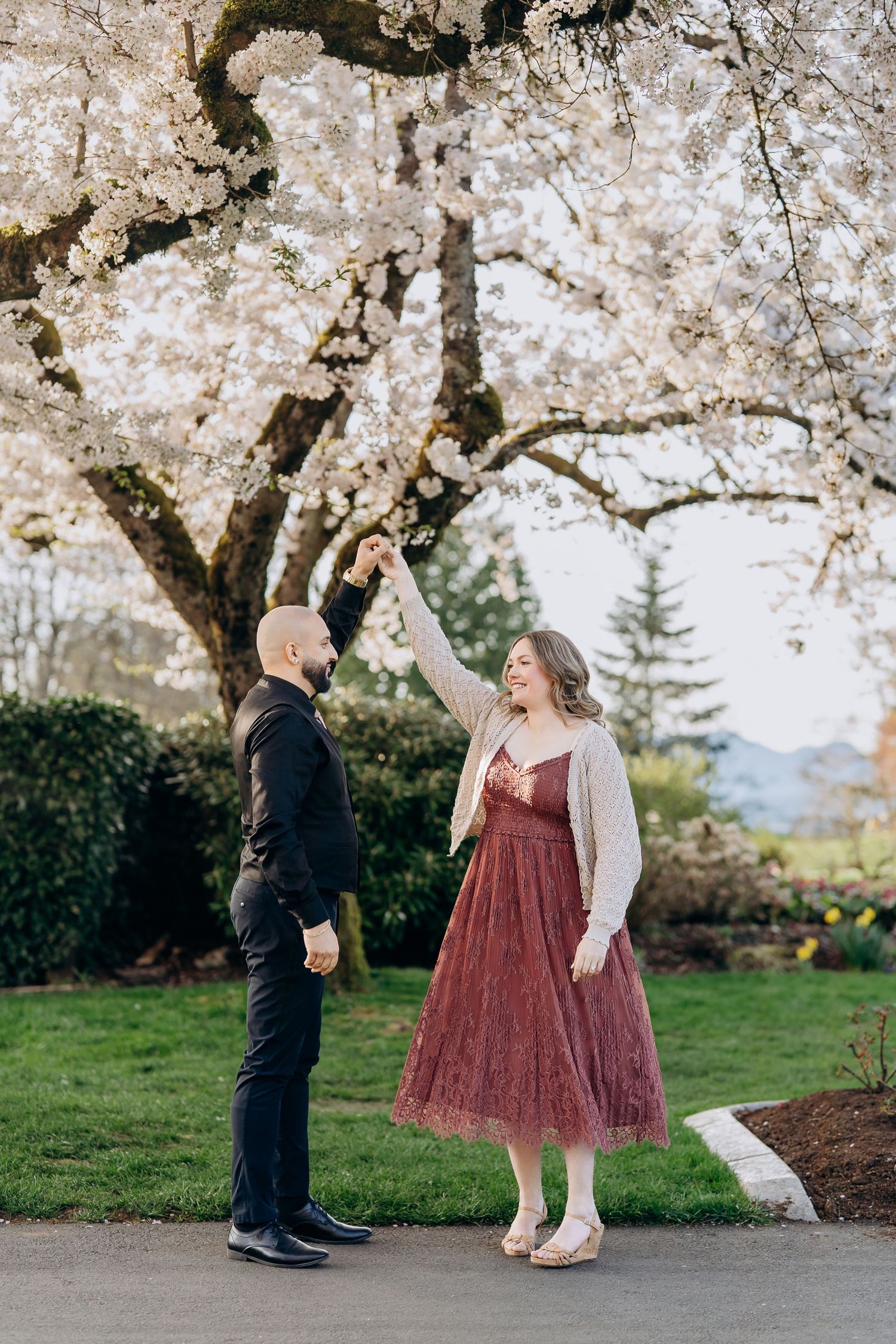 A romantic spring wedding scene unfolds beneath blooming cherry blossom trees with a burgundy dress and black suit.