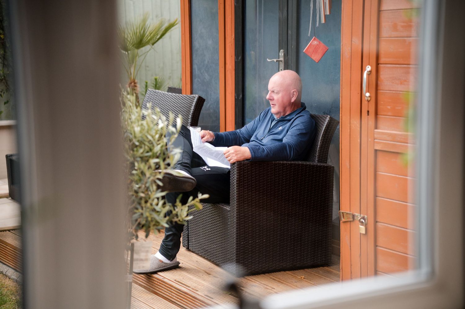 A person in a blue shirt sits on a porch swing while reading a book in front of a brick house.