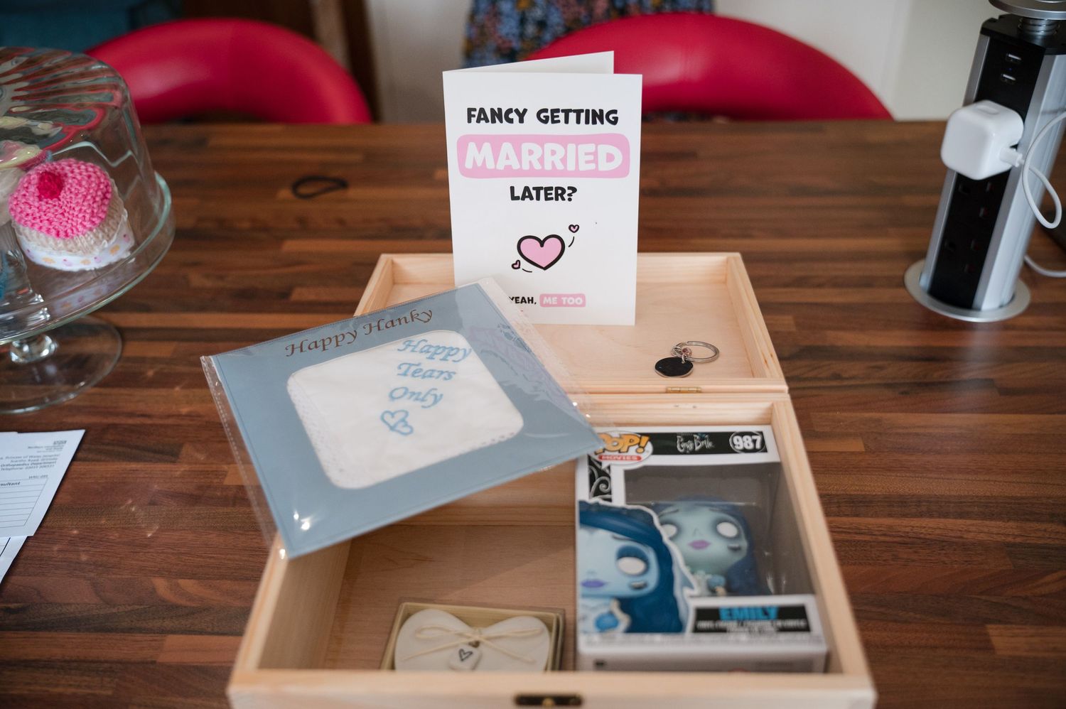 A wooden box containing wedding rings and cards sits on a table with a pink sign reading 'fancy getting married'.