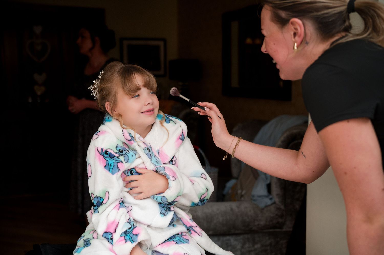 A person in a white floral robe is having makeup applied by someone wearing black.