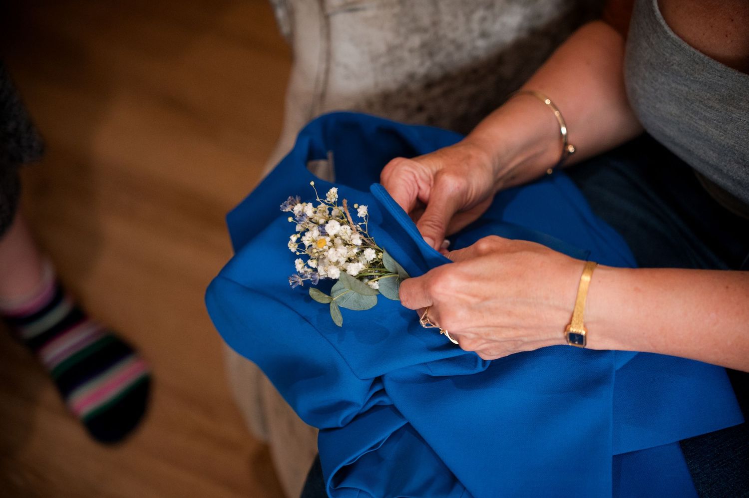 Hands delicately handle a small floral boutonniere against a bright blue fabric background.