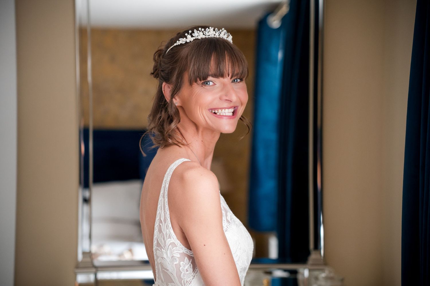 A bride in a white lace wedding dress and tiara smiles brightly while getting ready.