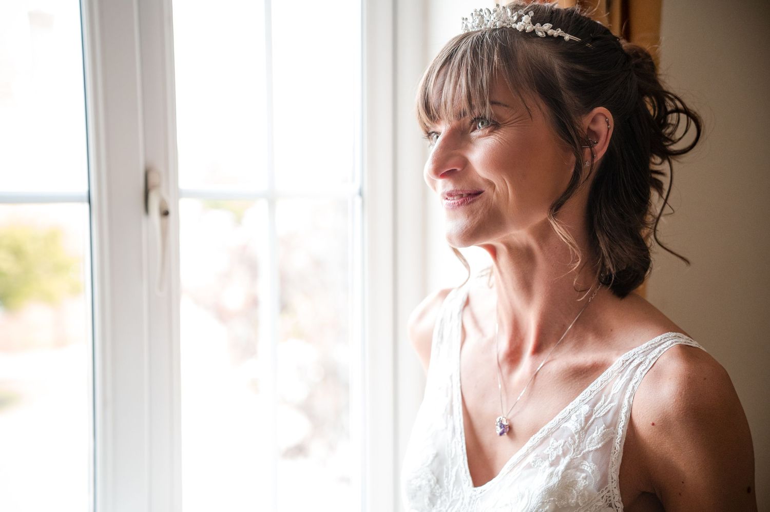 A person in a white lace dress gazes thoughtfully out a sunlit window.