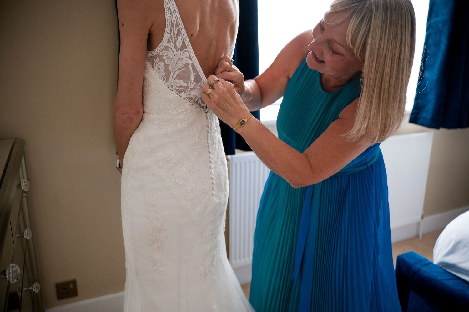 A person in a teal dress helps adjust a white wedding gown during a fitting session.