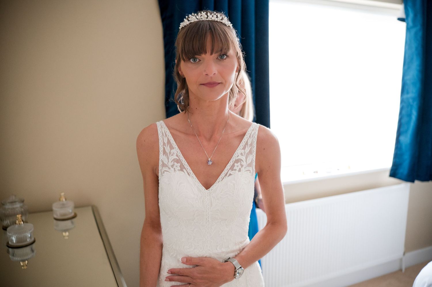 A bride in a white wedding dress sits at a desk writing in a journal near a window with blue curtains.