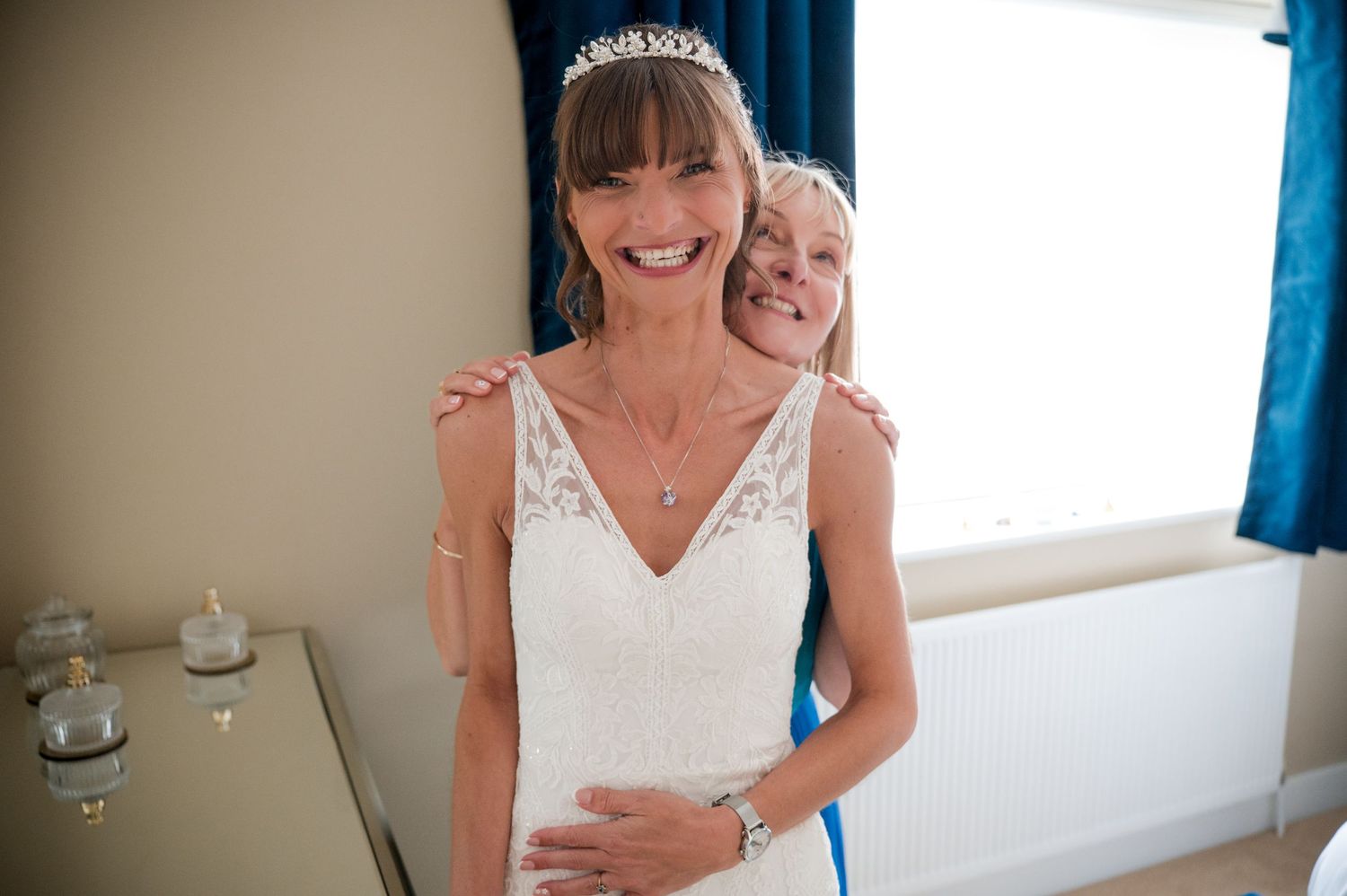 A bride in a white wedding dress smiles while getting ready in a room with blue curtains.
