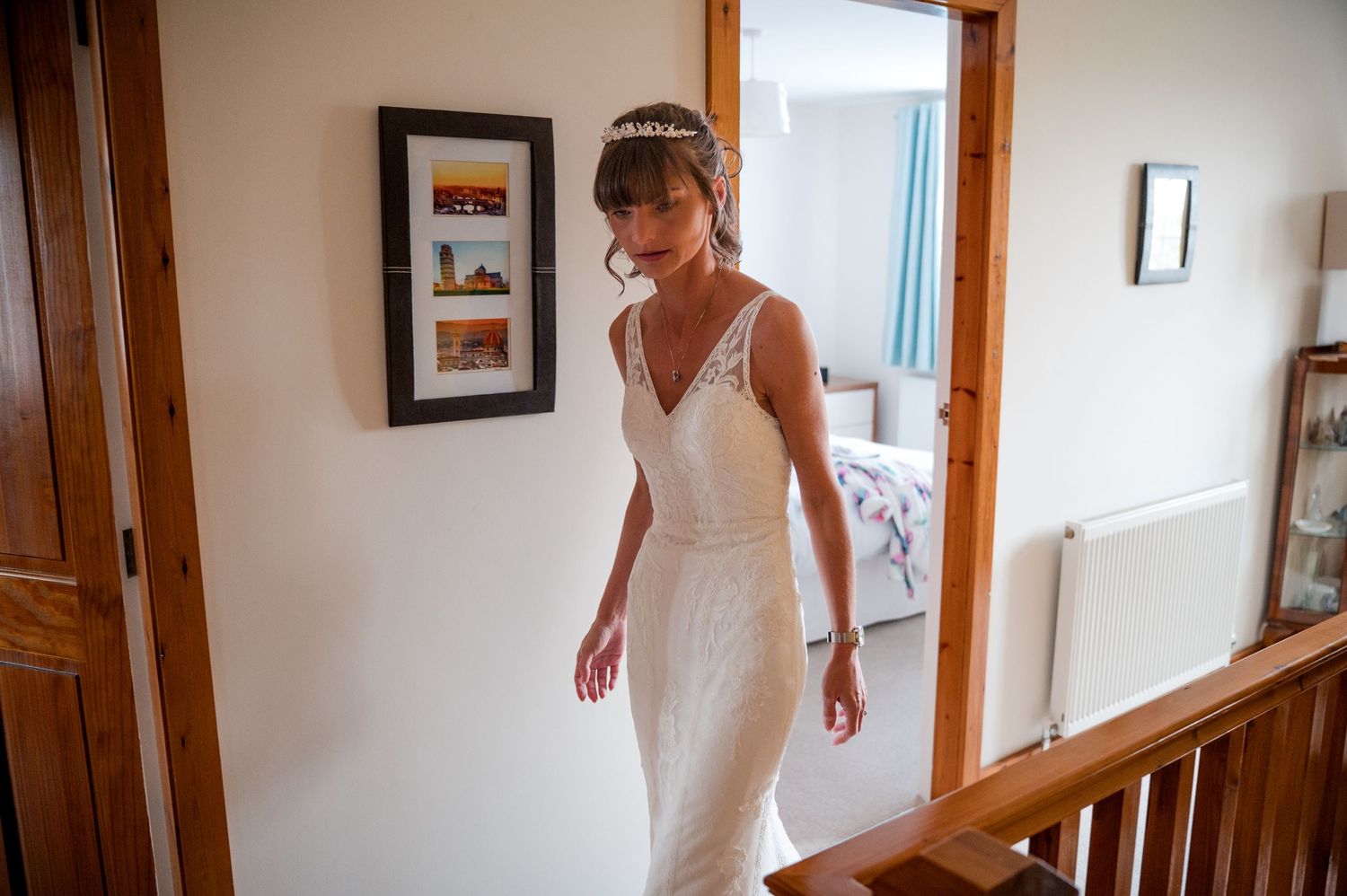 A bride in a white wedding dress walks down a wooden staircase in a sunlit home interior.