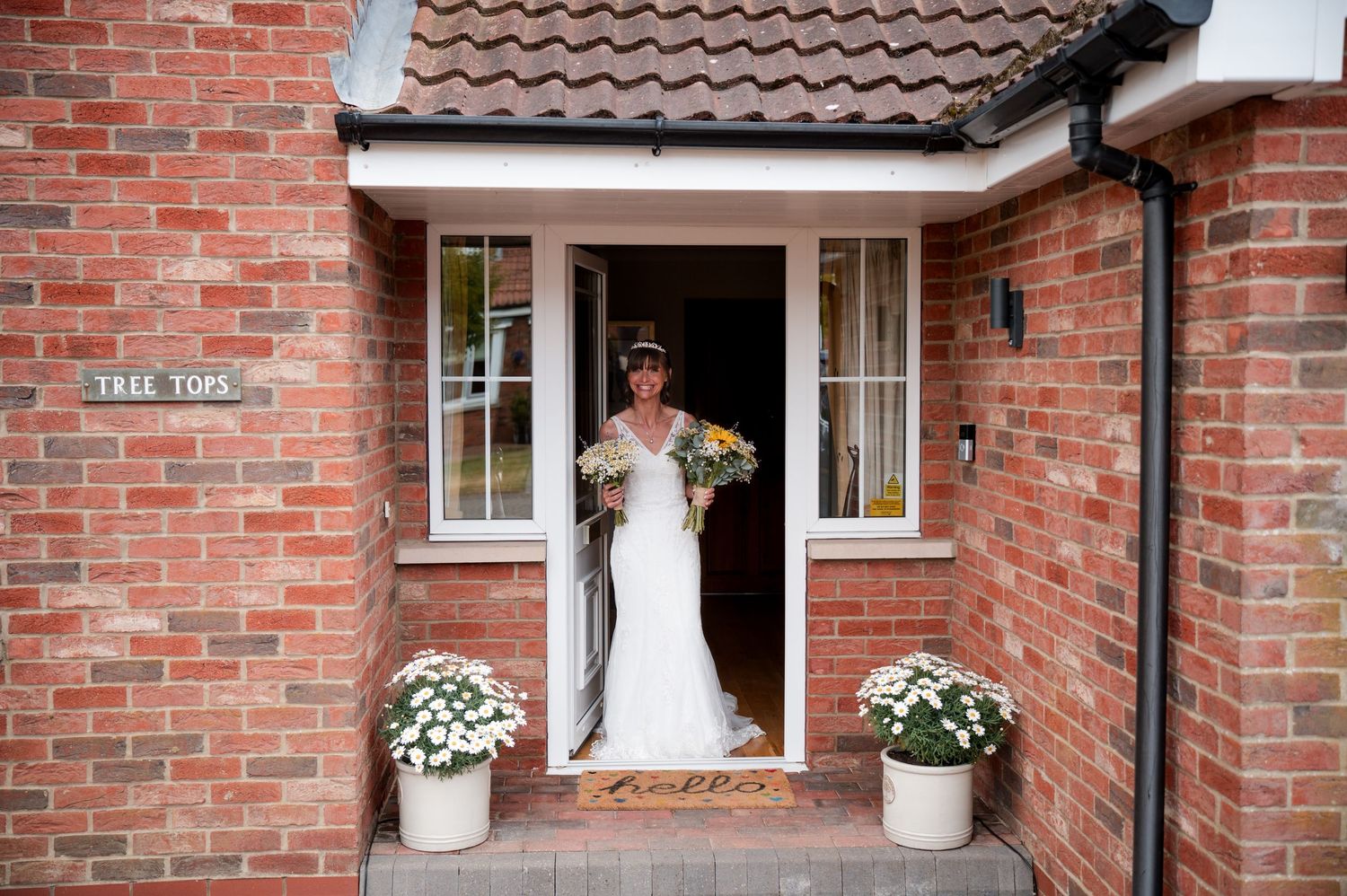 A bride in a white wedding dress poses in front of a red brick house with white trim and decorative flowers.