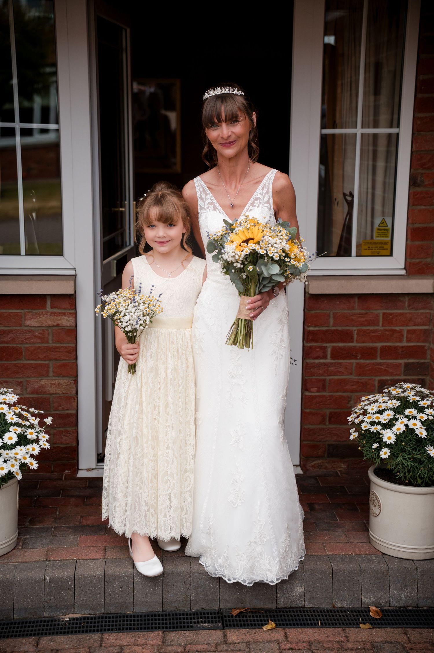A bride in a white wedding gown stands with a young flower girl in a cream lace dress holding bouquets.