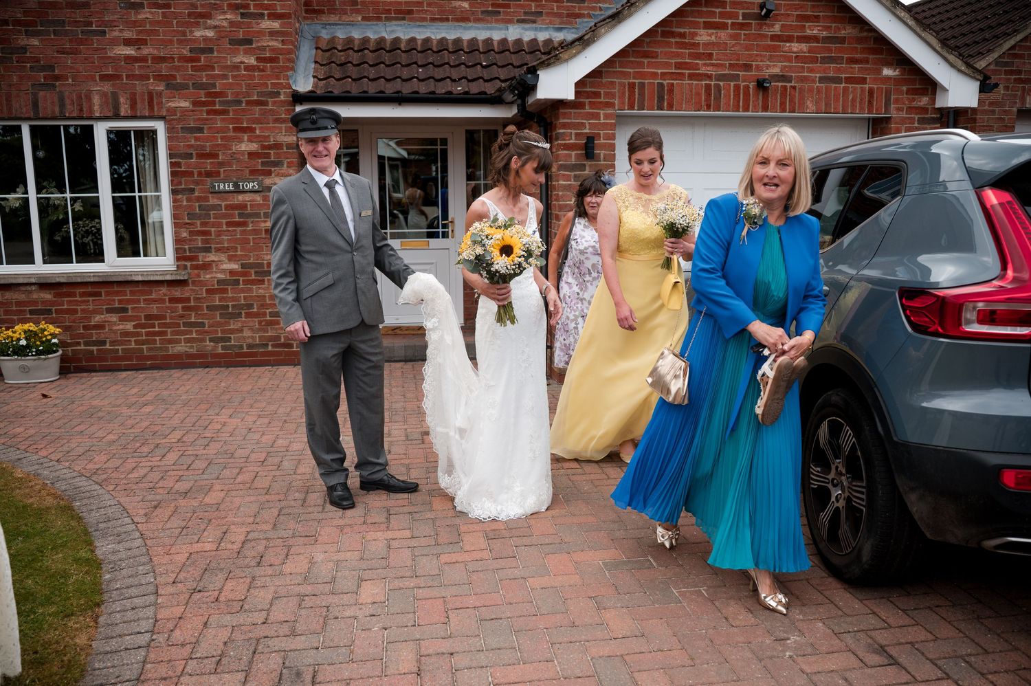 A group gathers outside a brick building with cars parked on a paved driveway.