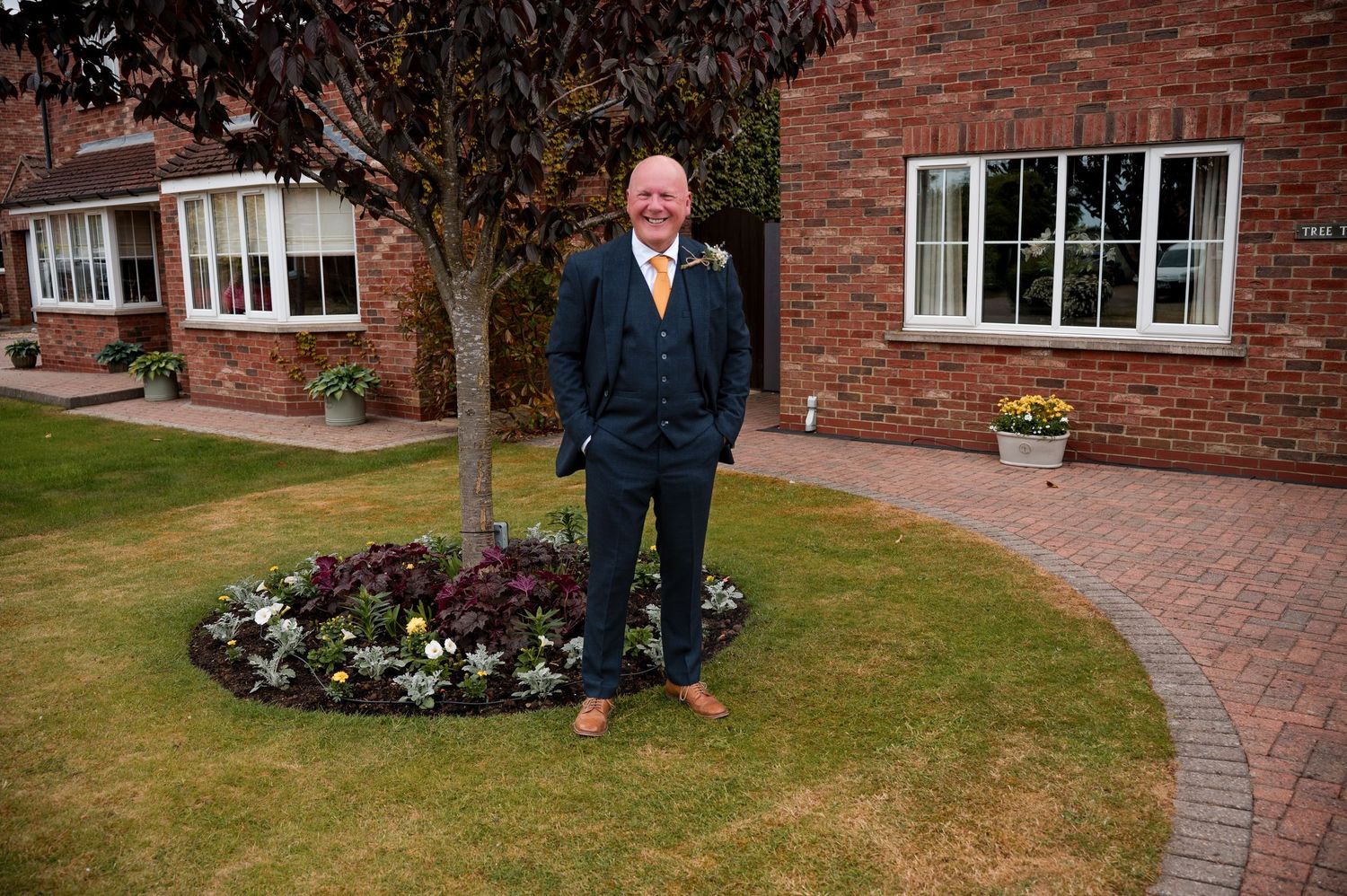 A person stands in front of a brick house next to a curved garden path and flowerbed on a manicured lawn.