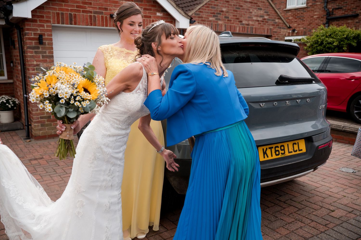 People in formal attire including blue and yellow dresses interact in front of a brick building.