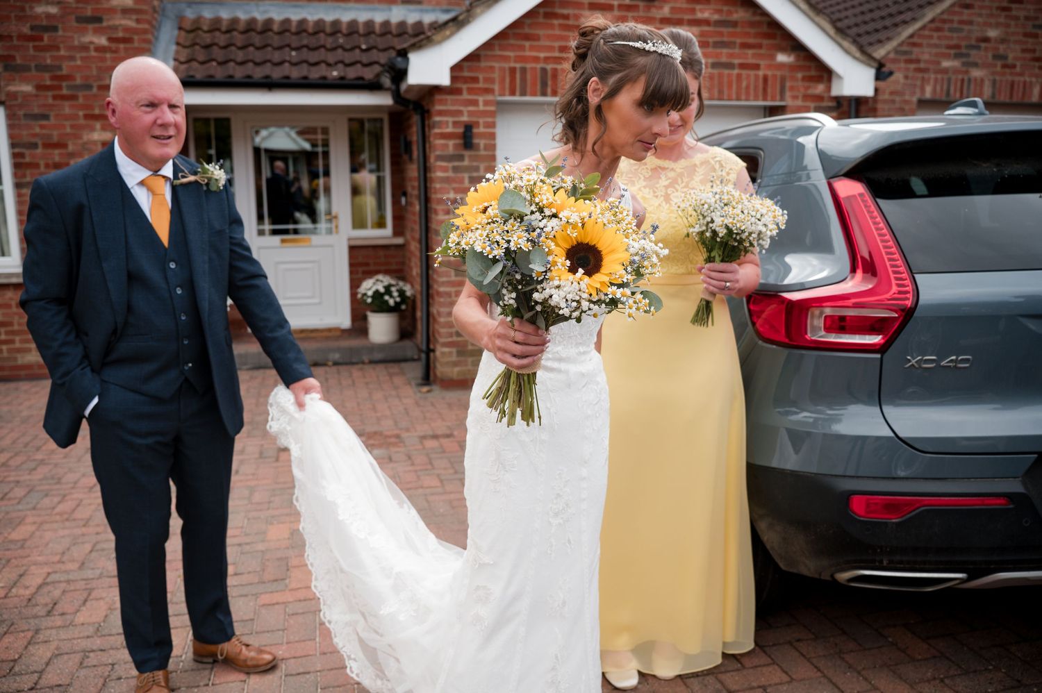 A wedding party walking down a brick path outside a building with a car parked nearby.