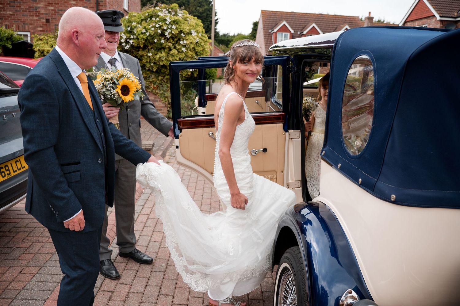 A wedding party arrives at a venue with guests helping the bride with her flowing white dress train.