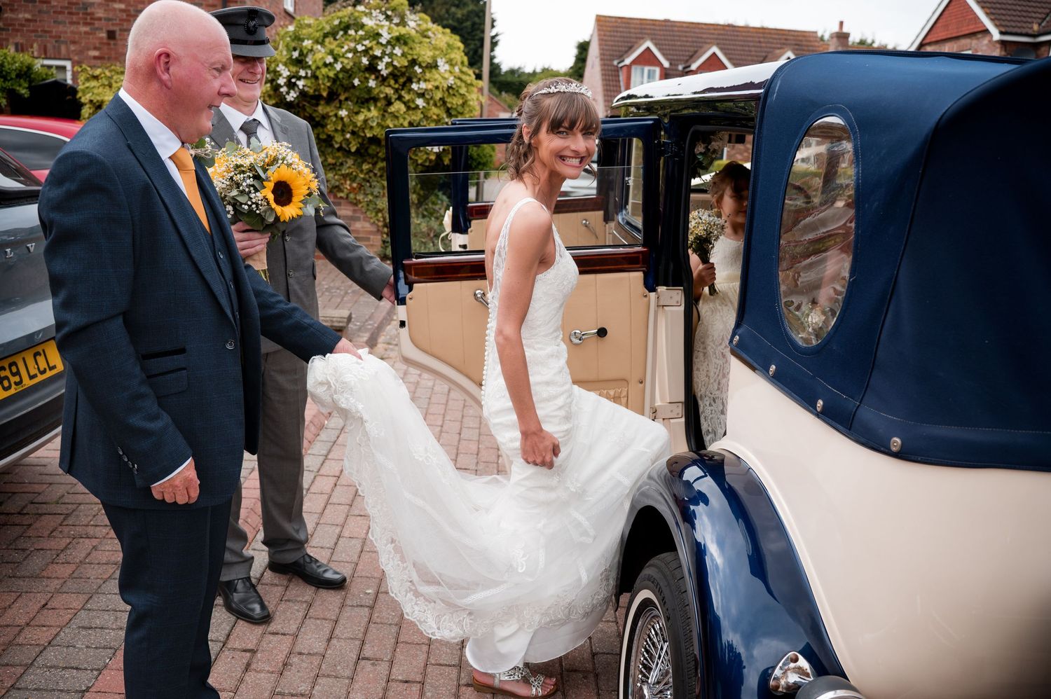 A bride in a white wedding dress steps into a vintage navy and cream colored classic car on a brick driveway.