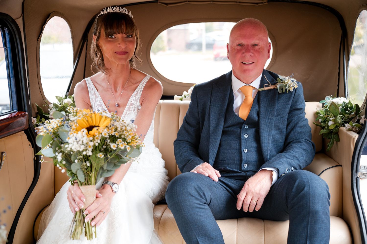 Newlywed couple sits in the back of a vintage car decorated with flowers for their wedding celebration.
