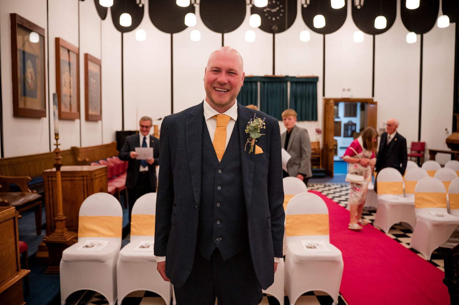 A person in a dark suit with yellow tie stands smiling in a wedding venue with white chair covers and red carpet.